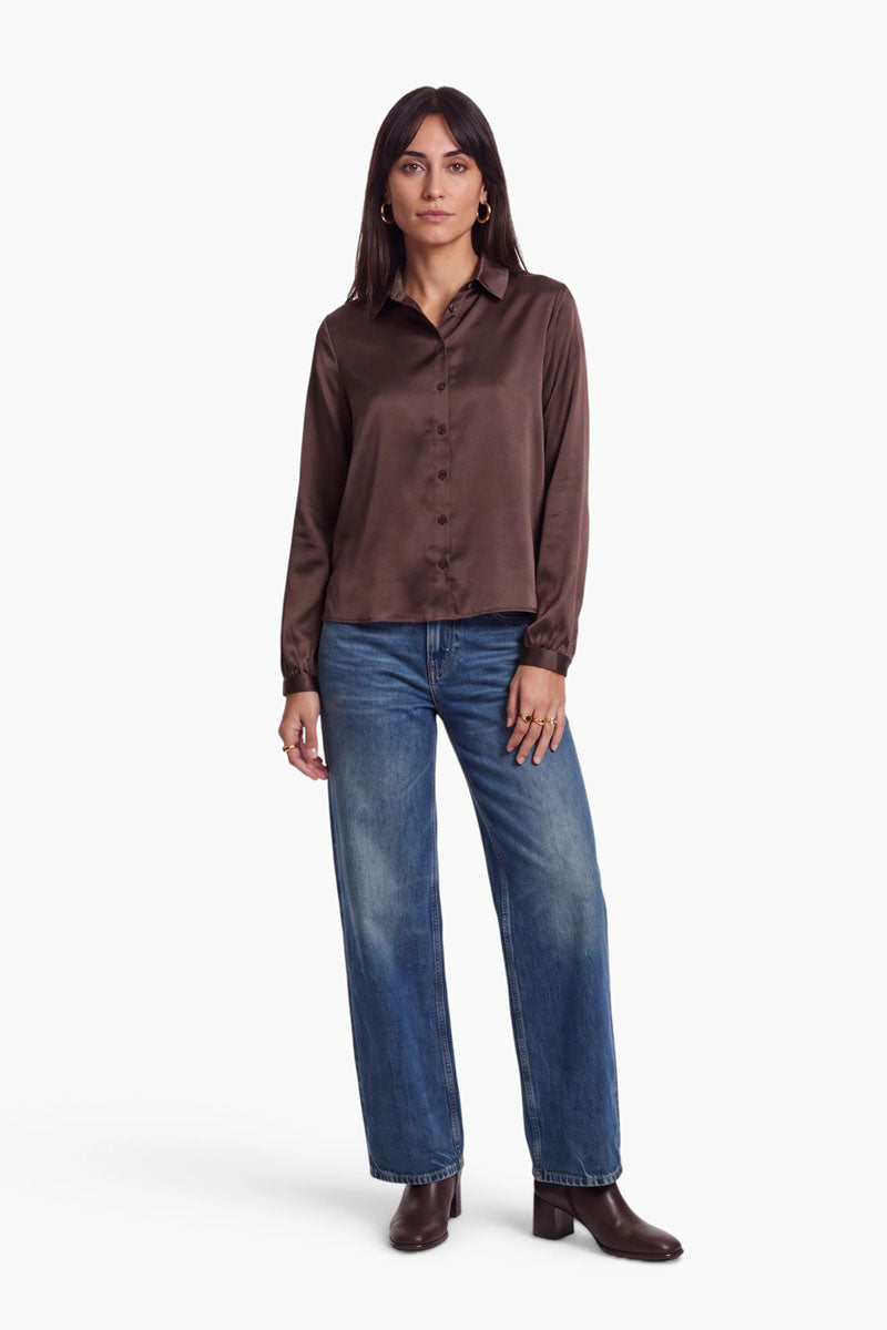 Eine Frau trägt eine langärmelige, braune Stretch-Bluse, die sie in eine mittelblaue, gerade geschnittene Bluejeans gesteckt hat. Sie steht und trägt braune Stiefel mit Blockabsatz.