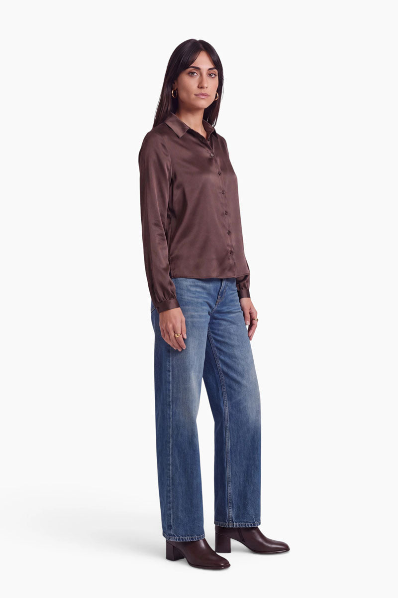 Eine Frau, die eine dunkelbraune Stretch-Bluse und eine blaue Jeans mit weitem Bein trägt. Sie steht und blickt leicht nach links; dazu trägt sie passende dunkelbraune Stiefeletten.