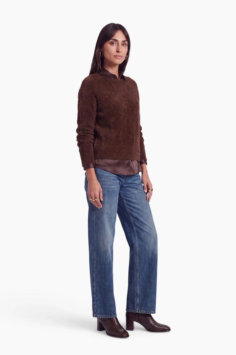 Eine Frau, die einen braunen Pullover mit Rundhalsausschnitt über einer passenden Stretch-Seidenbluse und einer blauen Jeans trägt. Sie trägt dunkelbraune Stiefeletten und goldene Ringe.