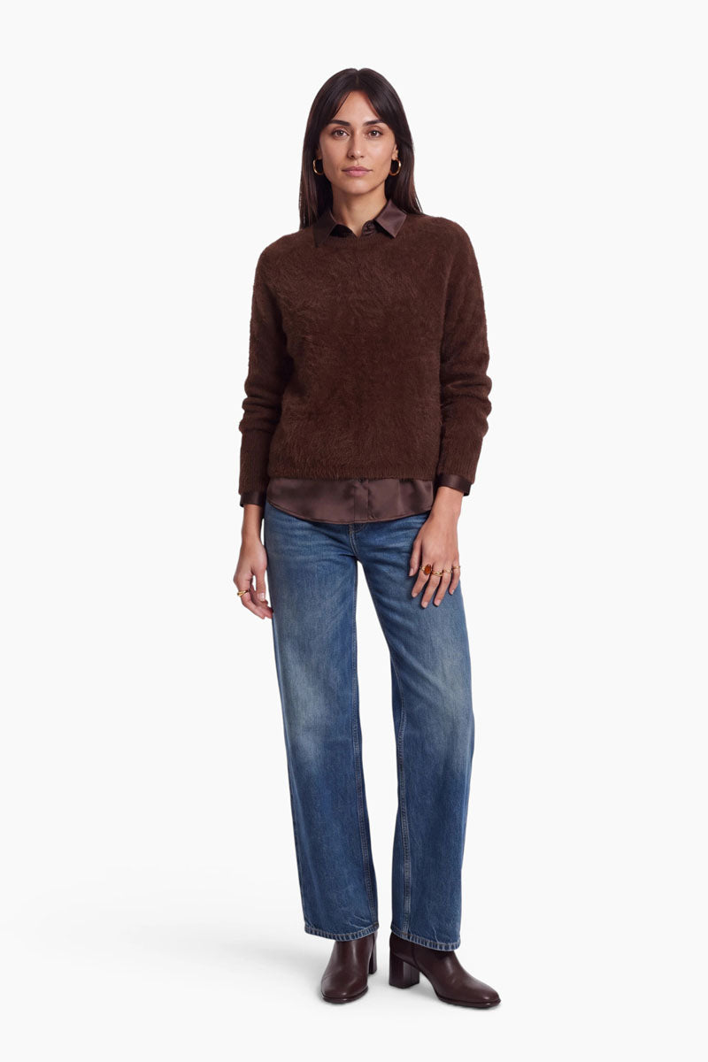 Eine Frau, die einen braunen, flauschigen Pullover über einer passenden Hemdbluse mit Knöpfen und einer blauen Jeans trägt. Sie steht und blickt in die Kamera; sie trägt goldene Ringe und dunkelbraune Stiefel mit Absatz.