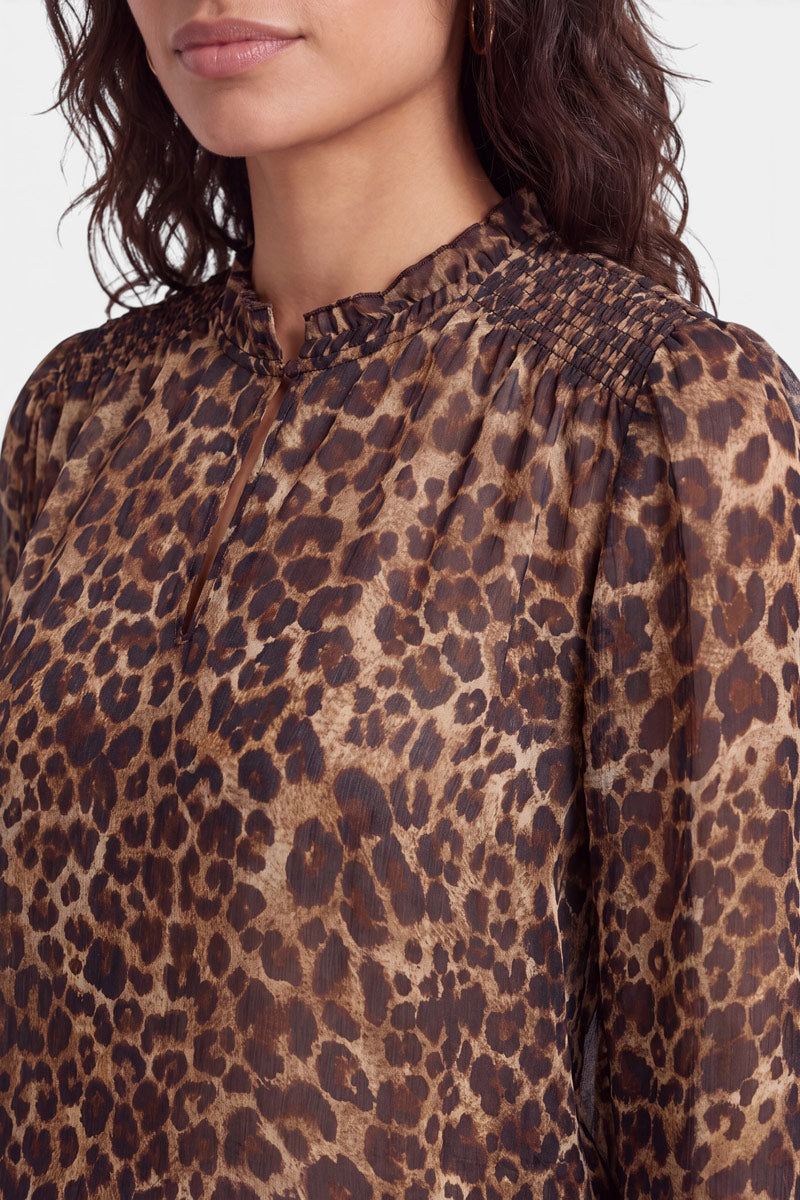 Eine Frau trägt eine durchsichtige, langärmelige Bluse mit Leopardenmuster in Braun und Schwarz. Die Bluse hat einen gerüschten Stehkragen, einen kleinen Schlitz vorne, geraffte Schultern, geraffte Ärmelbündchen und eine lockere Passform. Zu sehen sind nur ihr unterer Gesichtsbereich und ihr oberer Oberkörper.