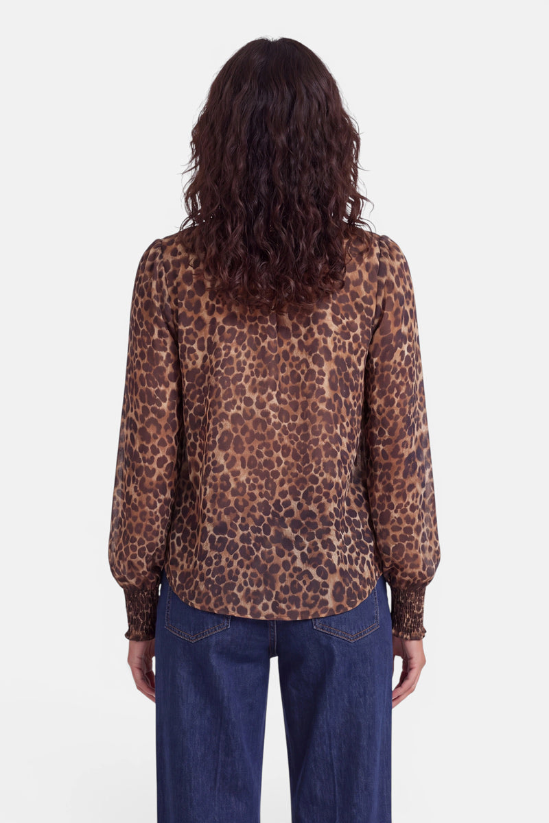 Eine Frau mit welligem braunem Haar steht mit dem Rücken zur Kamera. Sie trägt eine langärmelige Bluse mit Leopardenmuster und Smok-Bund, die über geraffte Ärmelbündchen und einen Stehkragen verfügt, dazu eine lockere blaue Jeans. Der Hintergrund ist schlicht weiß.