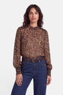 Eine Frau mit welligem, dunklem Haar trägt eine langärmelige Bluse mit Leopardenmuster, Smok-Bund und Stehkragen, kombiniert mit einer blau-hoch taillierten Jeans und einem braunen Gürtel. Sie steht vor einem schlichten weißen Hintergrund, blickt nach vorne und hat einen entspannten Gesichtsausdruck.