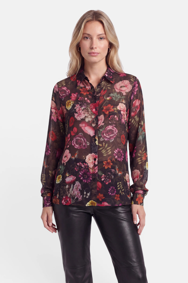 Eine Frau mit langen blonden Haaren trägt eine durchsichtige, schwarze, langärmelige Bluse mit einem lebhaften, bunten Blumenprint und eine schwarze Lederhose. Vor einem schlichten weißen Hintergrund blickt sie mit ausdruckslosem Gesicht nach vorne.