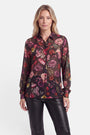 Eine Frau mit langen blonden Haaren trägt eine durchsichtige, schwarze, langärmelige Bluse mit einem lebhaften, bunten Blumenprint und eine schwarze Lederhose. Vor einem schlichten weißen Hintergrund blickt sie mit ausdruckslosem Gesicht nach vorne.