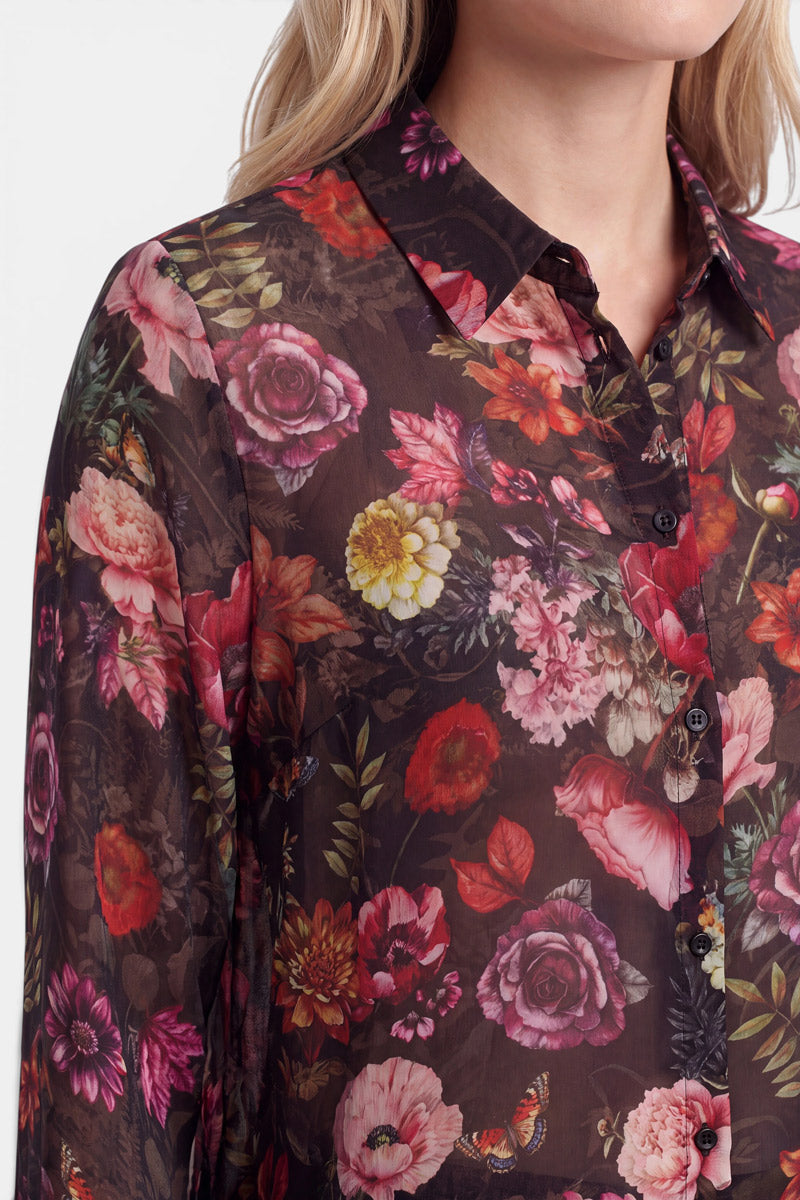 Eine Person mit hellem Haar trägt eine hauchdünne, transparente Bluse mit einem lebhaften, detailreichen Blumenprint in Rot-, Rosa- und Gelbtönen sowie grünen Blättern. Die Bluse hat einen Kragen und schwarze Knöpfe. Das Bild ist am Kinn und in der Mitte des Arms beschnitten.