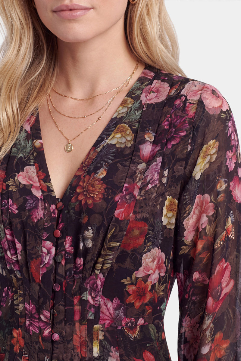 Eine Frau trägt ein transparentes, tailliertes, dunkles Kleid mit Blumenmuster, das mit rosa, roten und gelben Blumen verziert ist. Es handelt sich um ein Minikleid mit Blumenprint, Stoffknöpfen und einem V-Ausschnitt. Sie hat langes, welliges blondes Haar und trägt mehrere goldene Halsketten übereinander. Der Hintergrund ist schlicht weiß.