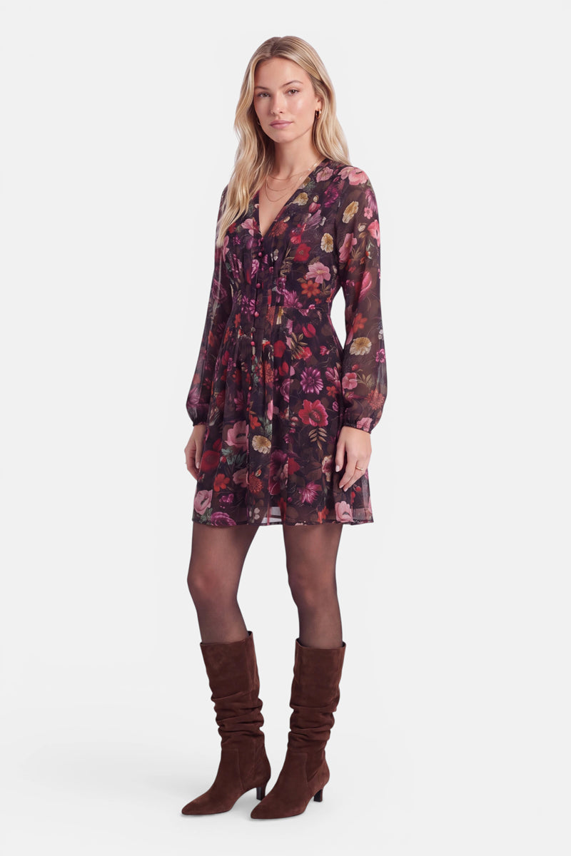 Eine Frau steht vor einem schlichten weißen Hintergrund und trägt ein langärmeliges Kleid mit dunklem Blumenmuster, schwarze, durchsichtige Strumpfhosen und braune, locker sitzende Stiefel, die bis zum Knie reichen. Sie hat blondes Haar, trägt nur wenig Schmuck und hat einen neutralen Gesichtsausdruck.