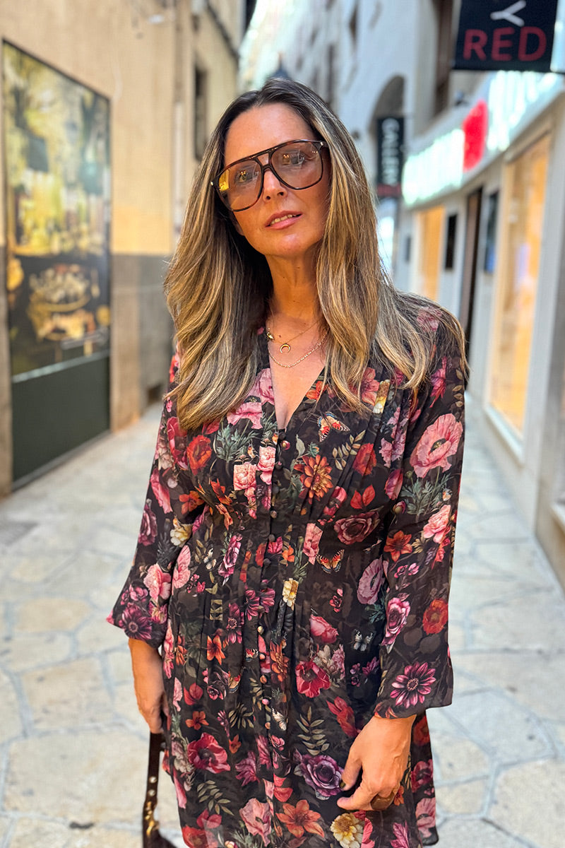 Eine Frau mit langen blonden Haaren, die eine große Brille und ein geblümtes Minikleid mit V-Ausschnitt trägt, steht auf einer gepflasterten Straße. Sie blickt mit ausdruckslosem Gesicht in die Kamera. Im unscharfen Hintergrund sind Geschäfte mit Leuchtreklamen und Kunstwerken in den Schaufenstern zu sehen.