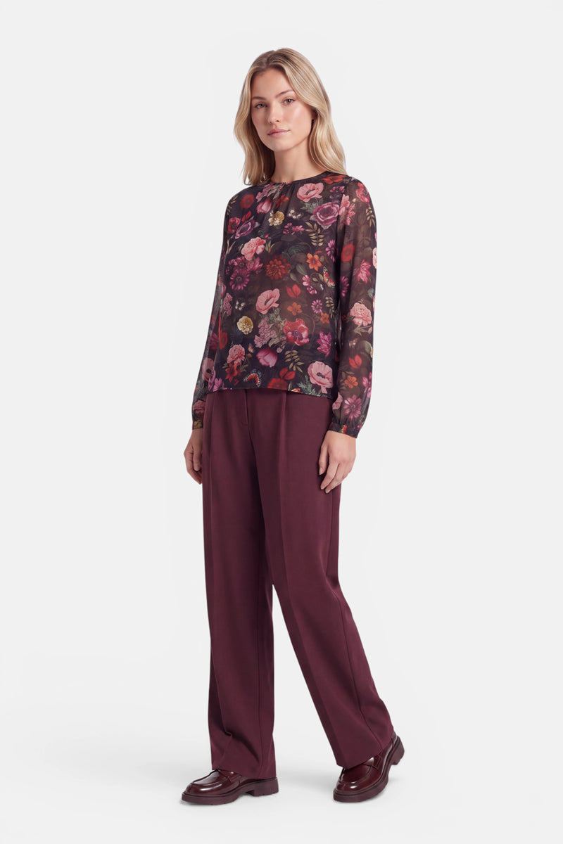 Vor einem weißen Hintergrund steht eine Frau, die eine langärmelige, durchsichtige Bluse mit rosa Blumenmuster trägt, dazu eine weite, burgunderrote Hose und dunkelbraune Schuhe. Sie hat helles Haar mit Mittelscheitel und einen neutralen Gesichtsausdruck.