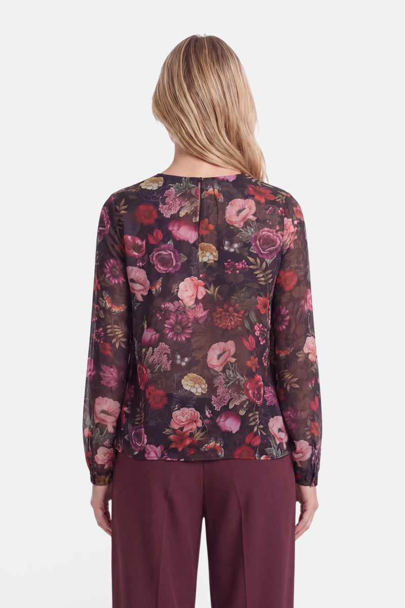 Eine Frau mit hellbraunen Haaren, von hinten gesehen, trägt eine durchsichtige, langärmelige Bluse mit einem bunten Blumenmuster und eine weinrote Hose. Auf der Bluse sind verschiedene rosa, rote und violette Blumen auf dunklem Hintergrund abgebildet.