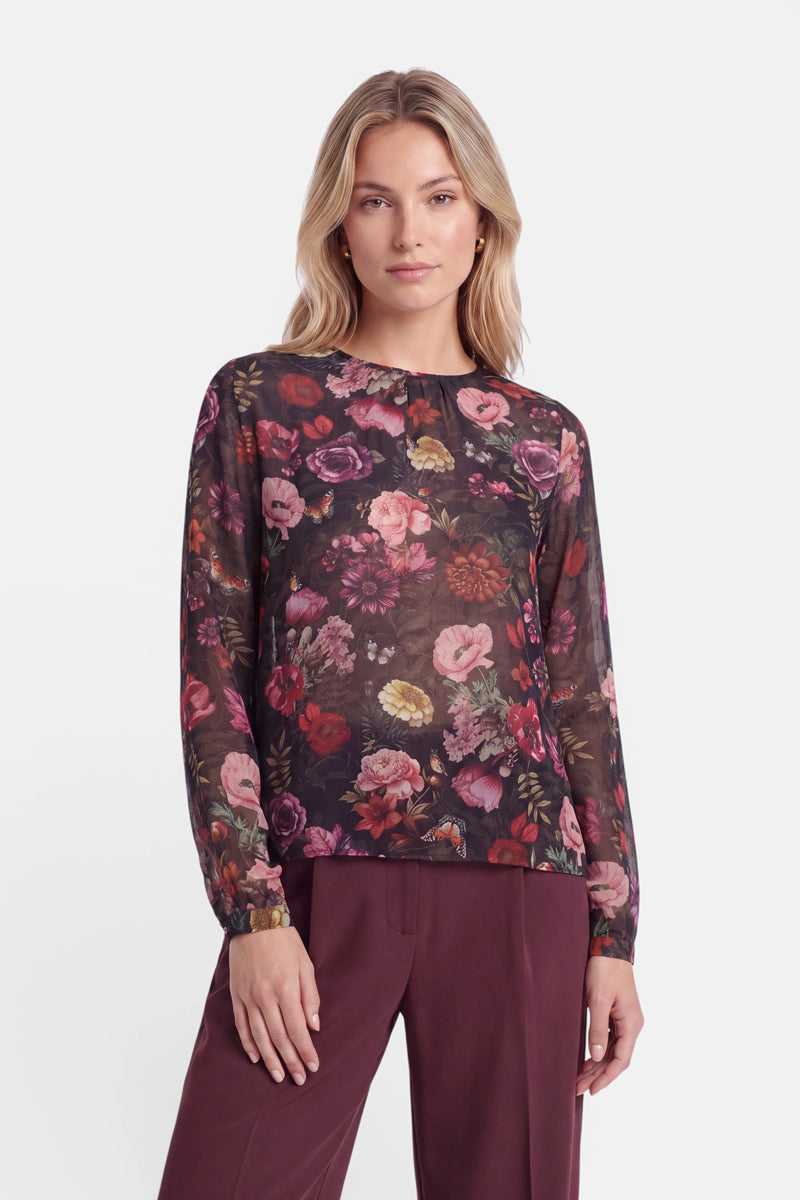Eine Person mit hellem Haar trägt eine durchsichtige, langärmelige Bluse mit einem lebhaften Blumenprint in Rot-, Rosa- und Violetttönen auf dunklem Hintergrund, dazu eine weinrote Hose, und steht vor einem schlichten weißen Hintergrund.