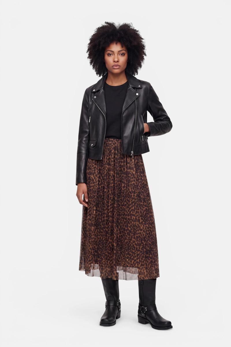 Eine Frau mit natürlich lockigem Haar trägt eine schwarze Lederjacke, ein schwarzes Oberteil und einen Midirock mit Leopardenmuster, dazu schwarze Stiefel. Sie steht mit neutralem Gesichtsausdruck vor einem schlichten weißen Hintergrund und blickt geradeaus.