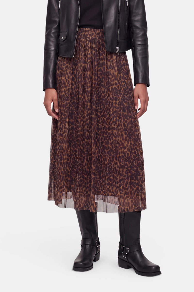 Eine Person, die eine schwarze Lederjacke, ein schwarzes Oberteil und einen braun-schwarzen Midirock mit Leopardenmuster trägt, dazu hohe schwarze Stiefel mit Schnallen, steht vor einem schlichten weißen Hintergrund. Das Foto ist unterhalb der Schultern beschnitten, sodass der Kopf nicht zu sehen ist.