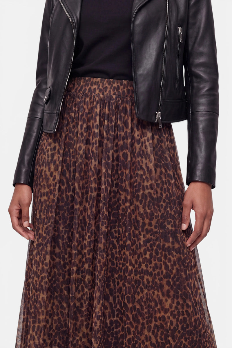 Eine Person trägt eine schwarze Lederjacke über einem schwarzen Oberteil und einen braunen Midirock mit Leopardenmuster und eleganten Falten. Das Foto ist vom Hals abwärts beschnitten und zeigt das Outfit vor einem schlichten, hellen Hintergrund.