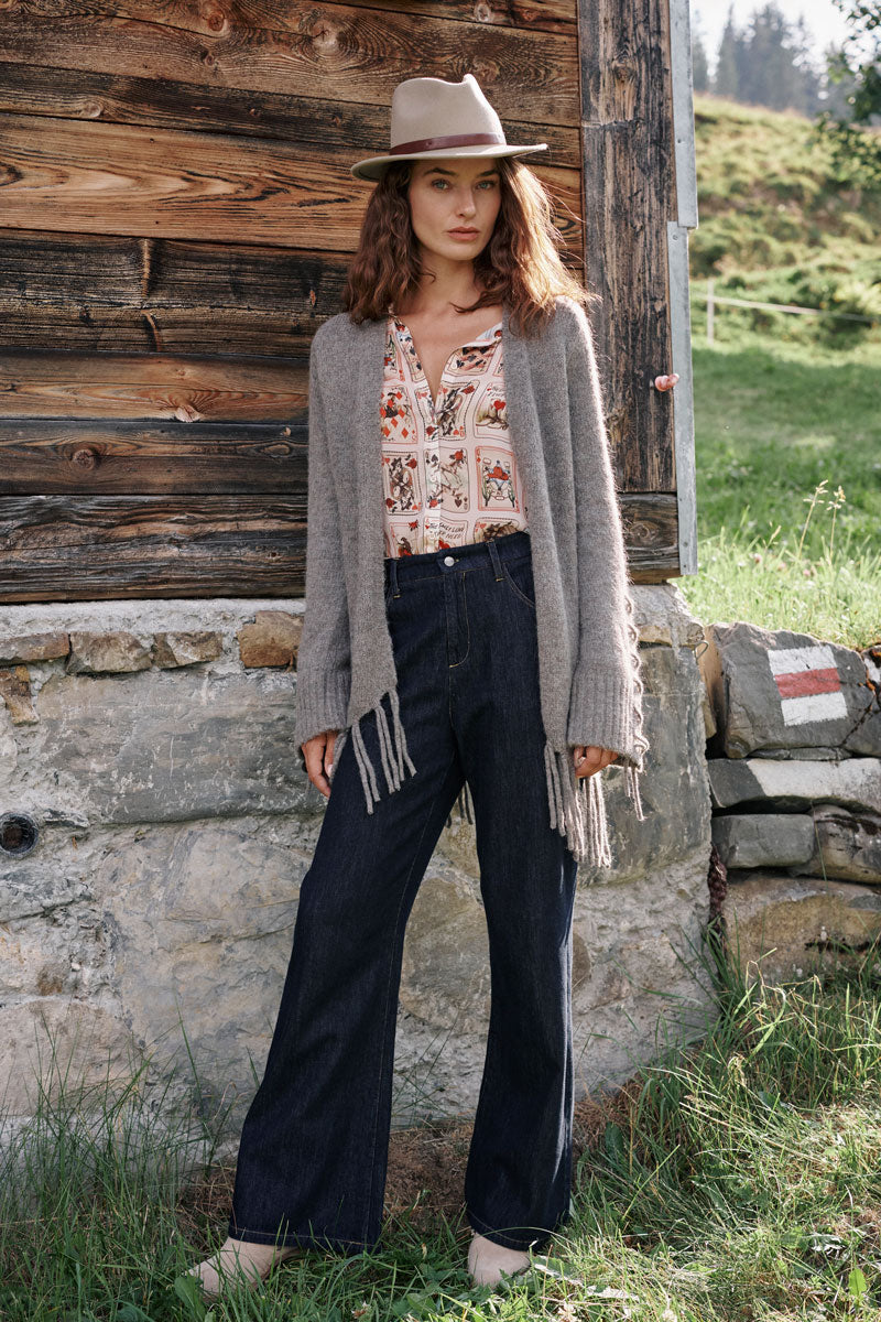 Eine Frau steht draußen vor einer Holzwand. Sie trägt einen hellbraunen Hut, eine graue Strickjacke aus einem Alpaka-Woll-Mix mit langen Fransen, eine gemusterte Bluse und eine dunkle Jeans mit weitem Bein. In der Nähe sind Gras und Steine mit roten und weißen Farbmarkierungen zu sehen, und im Hintergrund stehen Bäume.