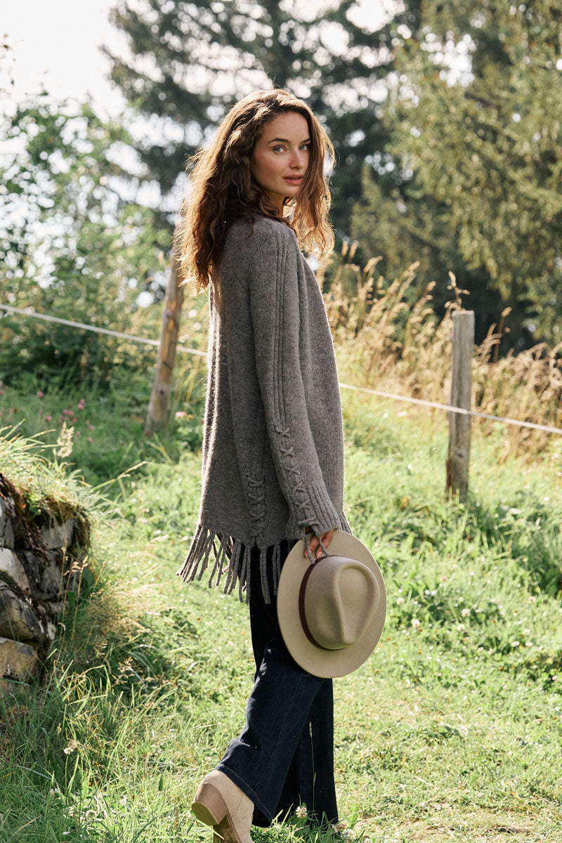 Eine Frau mit welligem braunem Haar steht draußen auf einer Wiese und trägt eine graue Strickjacke aus einem Alpaka-Woll-Mix mit langen Fransen, eine dunkle Jeans und beige Schuhe. Sie blickt über die Schulter zurück, während sie einen hellbraunen Hut in der Hand hält. Sonnenlicht fällt durch die Bäume und einen rustikalen Holzzaun.