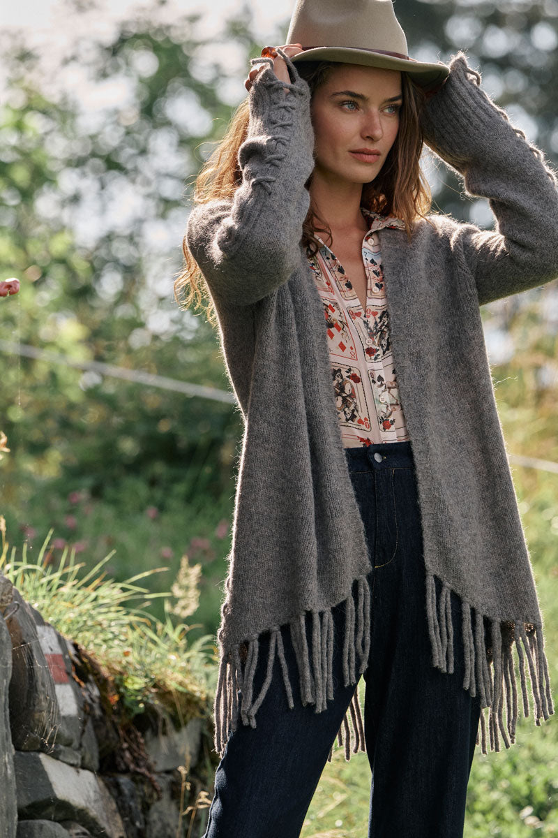 Eine Frau im Freien rückt ihren hellbraunen Hut zurecht. Sie trägt eine Strickjacke aus einem Alpaka-Woll-Mix mit langen Fransen über einer geblümten Bluse und einer dunkelblauen Jeans mit hoher Taille. Sie steht neben einer Steinmauer im sonnendurchfluteten Gras, im Hintergrund ist verschwommenes grünes Laub zu sehen.
