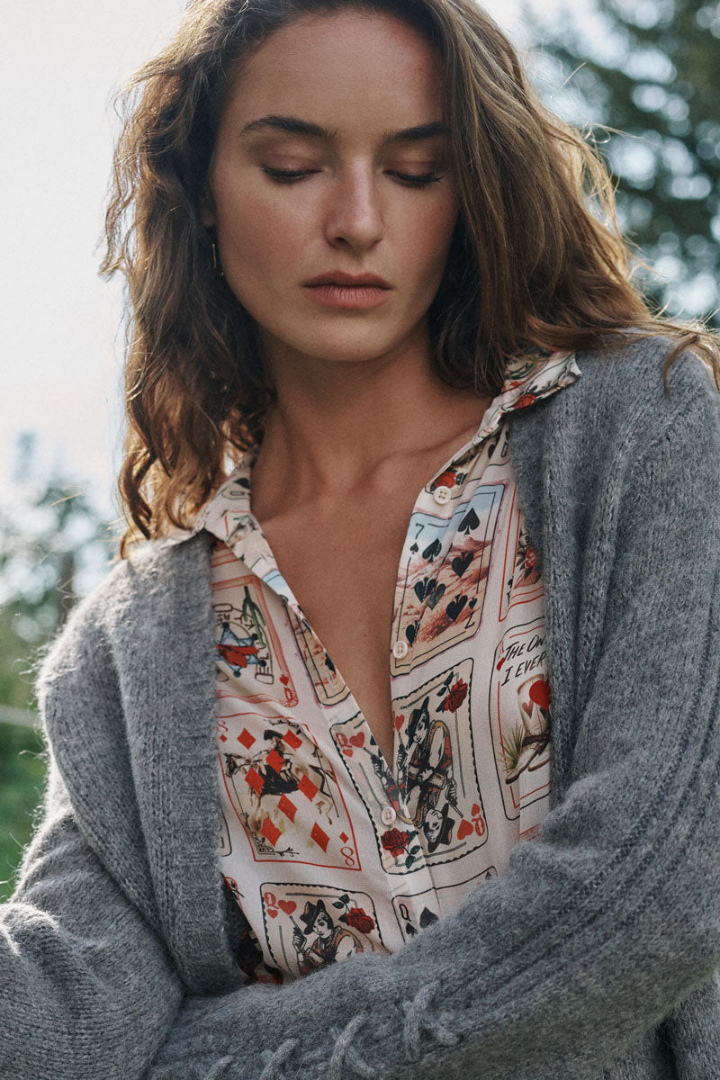 Eine Frau mit welligem braunem Haar blickt mit ausdruckslosem Gesicht nach unten. Sie trägt eine graue Strickjacke aus einem Alpaka-Woll-Mix über einer Hemdbluse, die mit Spielkartenmotiven verziert ist. Der Hintergrund besteht aus verschwommenem Grün, was auf eine Außenaufnahme hindeutet.