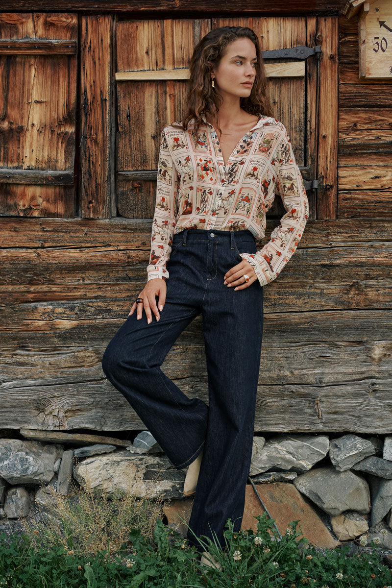 Eine Frau steht vor einer rustikalen Holzwand und trägt eine Hemdbluse mit einem von der Natur inspirierten Western-Karten-Print sowie eine dunkle Jeans mit weitem Bein. Sie blickt zur Seite, eine Hand in der Tasche, zu ihren Füßen sind Gras und Steine zu sehen.