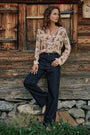 Eine Frau steht vor einer rustikalen Holzwand und trägt eine Hemdbluse mit einem von der Natur inspirierten Western-Karten-Print sowie eine dunkle Jeans mit weitem Bein. Sie blickt zur Seite, eine Hand in der Tasche, zu ihren Füßen sind Gras und Steine zu sehen.