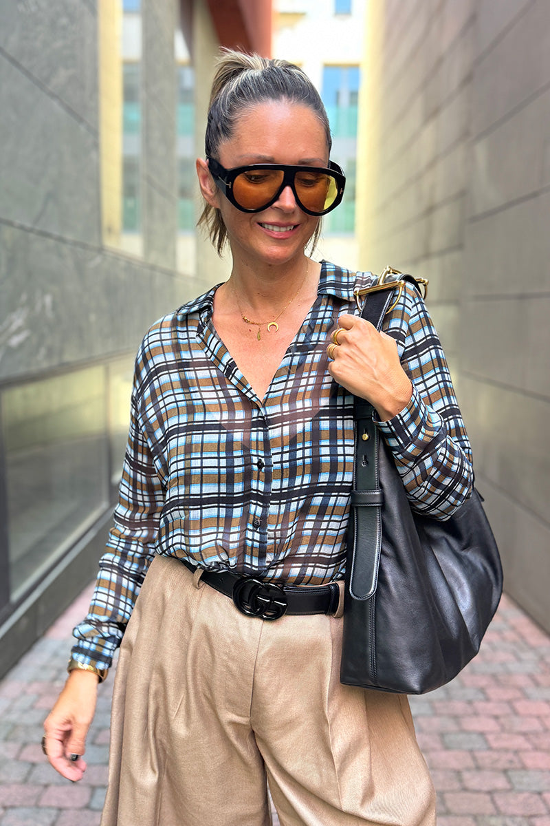 Eine Frau mit Pferdeschwanz lächelt in einer engen Gasse. Sie trägt eine übergroße Sonnenbrille, eine braune Hemdbluse mit Karoprint, eine beige Faltenhose mit schwarzem Gürtel und eine schwarze Umhängetasche. Die Sonne spiegelt sich im Fenster hinter ihr.