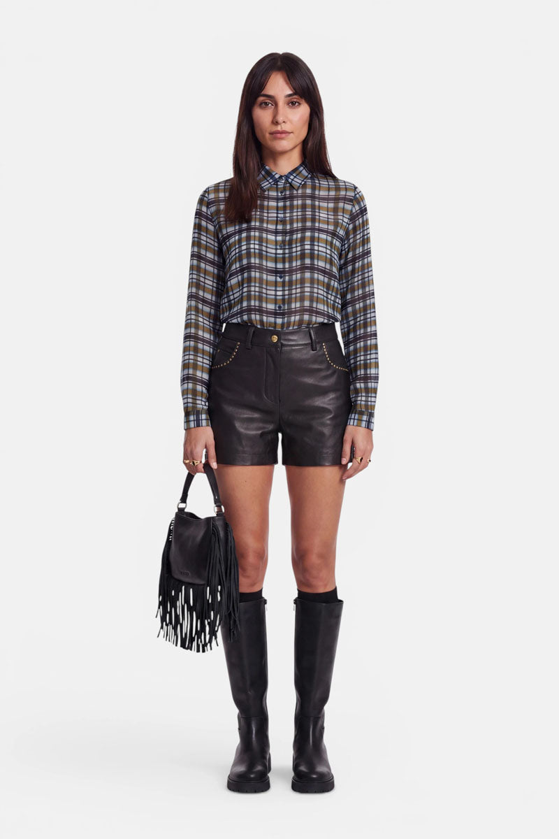 Eine Frau steht mit dem Blick nach vorne und trägt ein kariertes Hemd mit Knöpfen, das in schwarze Shorts aus veganem Leder mit goldenen Akzenten gesteckt ist. Sie hält eine schwarze Handtasche mit Fransen in der Hand und trägt schwarze kniehohe Stiefel. Der Hintergrund ist schlicht weiß.