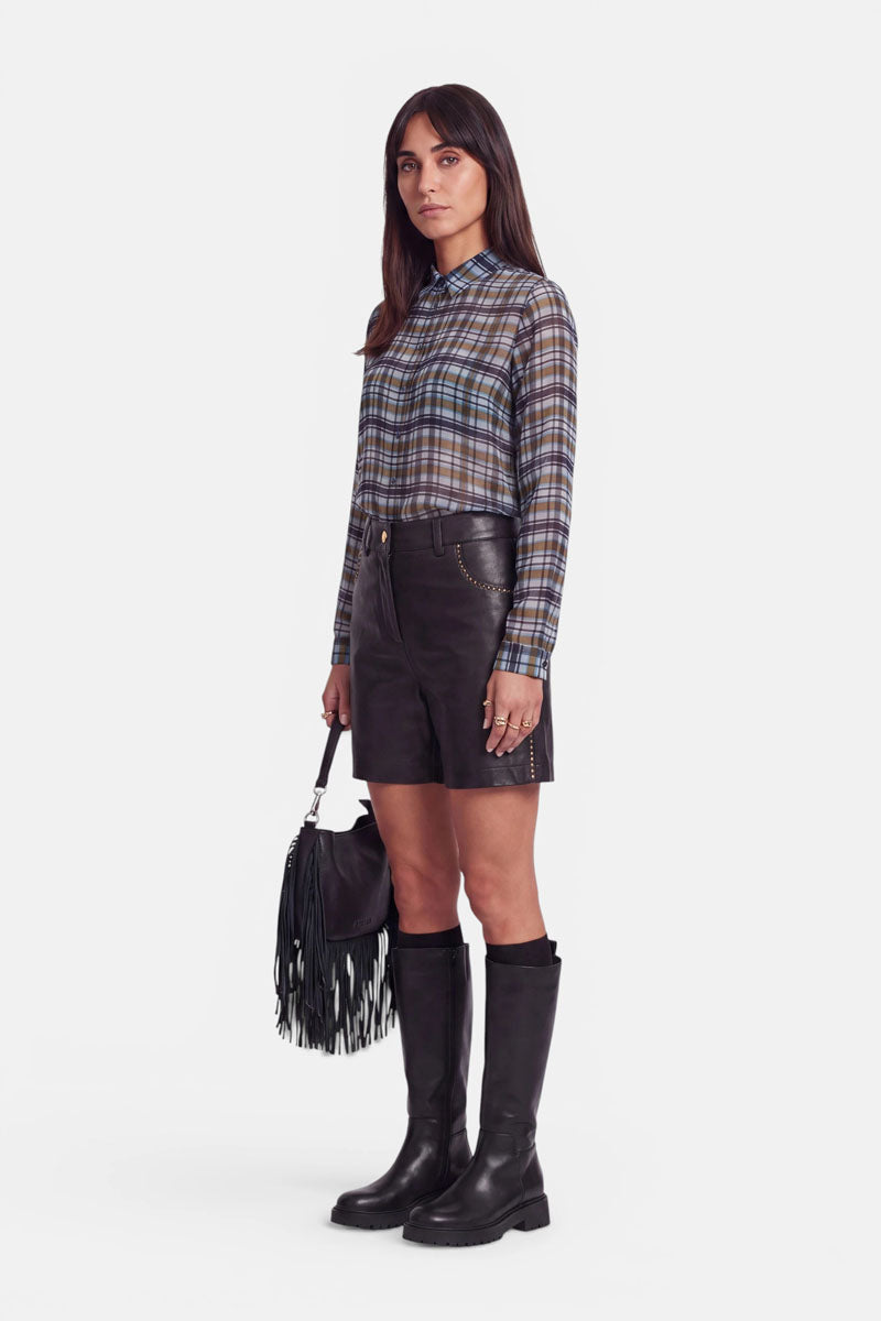 Eine Frau steht mit dem Blick nach vorne und trägt eine blaue, karierte Hemdbluse, die in schwarze Shorts mit hoher Taille und Nieten gesteckt ist, sowie schwarze kniehohe Stiefel; in der Hand hält sie eine schwarze Handtasche mit Fransen. Ihr glattes braunes Haar fällt ihr über die Schultern. Der Hintergrund ist schlicht weiß.