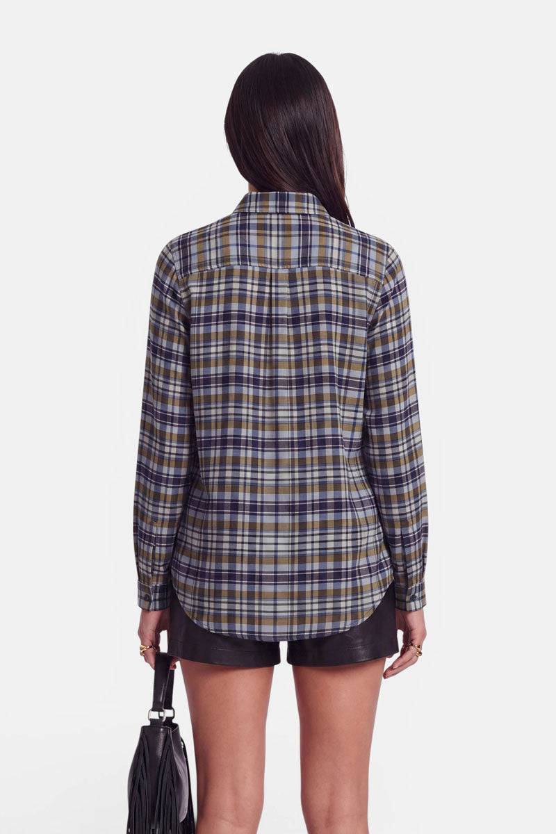 Eine Frau steht mit dem Rücken zur Kamera und trägt eine langärmelige Karoprint-Hemdbluse in Blau-, Gelb- und Weißtönen, dazu schwarze Ledershorts. In der linken Hand hält sie eine schwarze Tasche mit Fransen. Ihr glattes, dunkles Haar fällt vor einem weißen Hintergrund über ihren Rücken.
