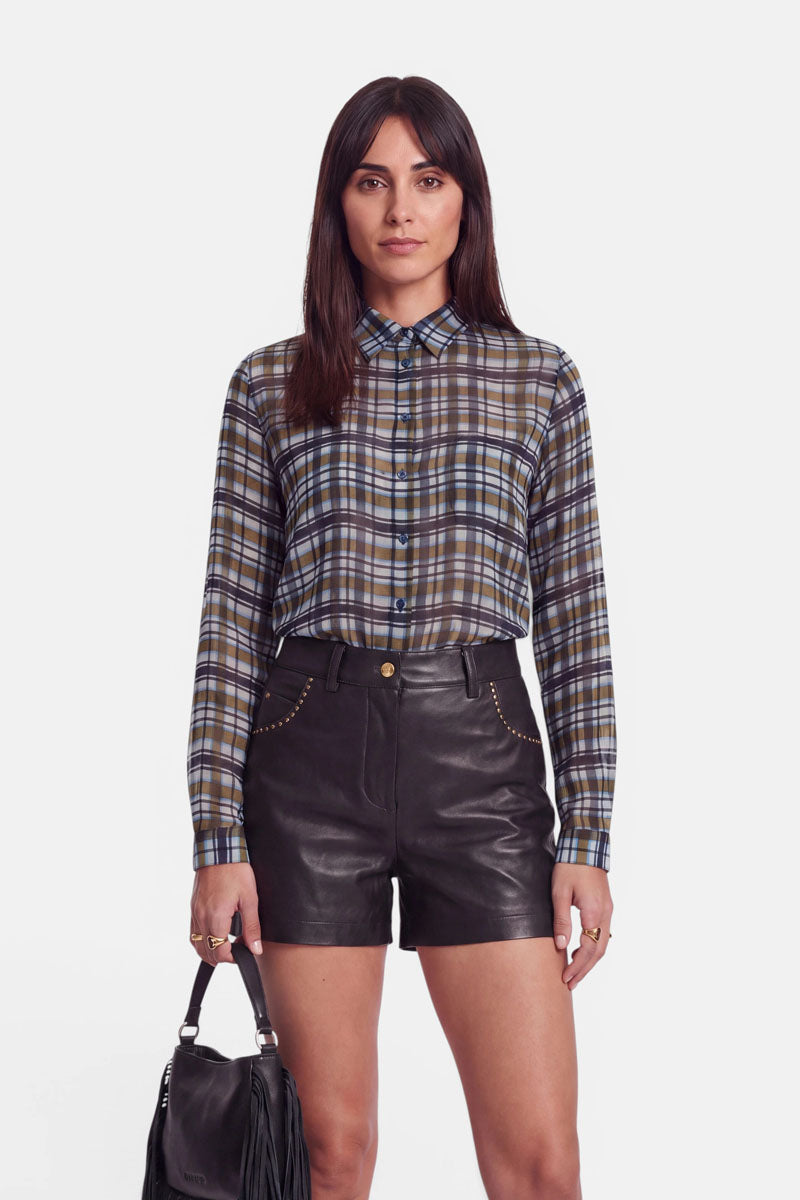 Eine Frau mit glattem braunem Haar trägt ein geknöpftes Karohemd und schwarze Shorts mit hoher Taille aus veganem Leder, die an den Taschen mit goldenen Nieten verziert sind. Sie hält eine schwarze Handtasche mit Fransen in der linken Hand und steht vor einem schlichten weißen Hintergrund, den Blick nach vorne gerichtet.