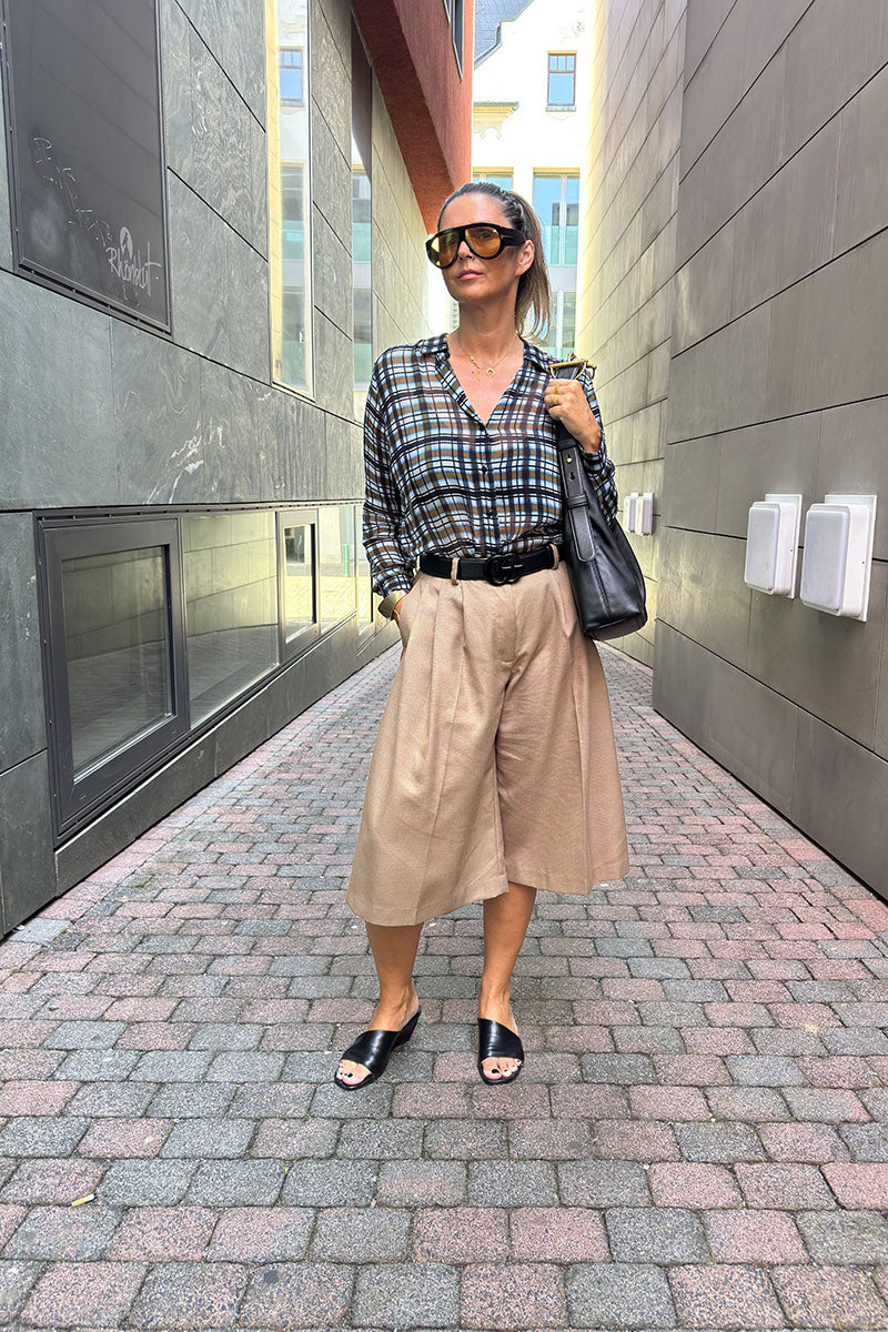 Eine Frau steht in einer städtischen Gasse. Sie trägt eine Sonnenbrille, eine Hemdbluse mit Karoprint, die in eine hellbraune Culotte gesteckt ist, schwarze Sandalen und eine schwarze Umhängetasche. Die Kulisse ist modern, mit hohen Gebäuden und Kopfsteinpflaster.