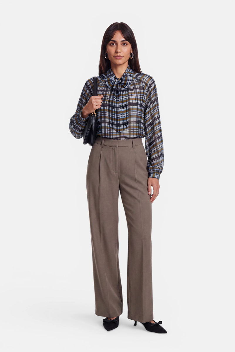 Eine Frau steht mit dem Blick nach vorne und trägt eine Karoprint-Bluse mit V-Ausschnitt und einer Schleife am Hals, eine taupefarbene Hose mit hoher Taille und weitem Bein sowie schwarze spitze Schuhe. Sie hält eine schwarze Umhängetasche in der Hand, hat glattes dunkles Haar und posiert vor einem schlichten hellen Hintergrund.