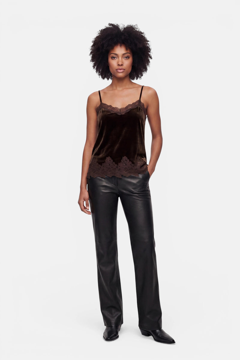 Eine Frau steht mit dem Blick nach vorne und trägt ein dunkelbraunes Samt-Top mit Spitzenbesatz, eine schwarze vegane Lederhose und schwarze Schuhe mit spitzer Zehenpartie. Sie hat einen neutralen Gesichtsausdruck, eine Hand in der Tasche und kurzes, lockiges Haar. Die gerade geschnittene Hose verleiht ihrem Look vor dem schlichten weißen Hintergrund eine moderne Note.