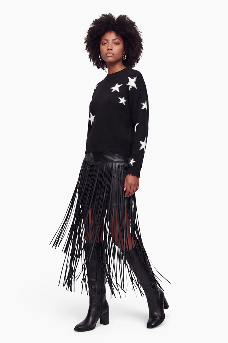 Eine Frau steht in einem schwarzen Pullover mit weißen Sternen, dazu trägt sie einen Minirock aus veganem Leder mit langen Fransen und kniehohe schwarze Stiefel. Sie hat eine natürliche Afro-Frisur und trägt Creolen, während sie selbstbewusst in die Kamera blickt. Der Hintergrund ist weiß.