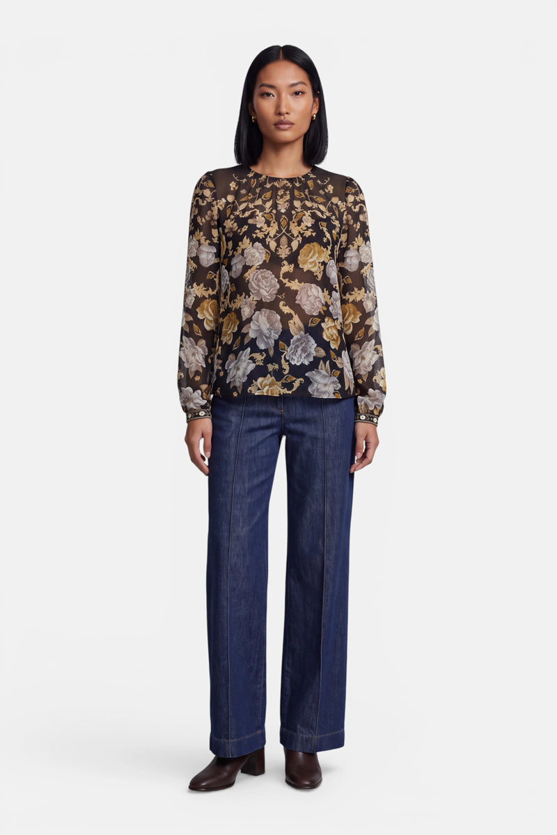 Eine Frau steht mit dem Blick nach vorne und trägt eine transparente Bluse mit einem gelben, grauen und taupefarbenen Blumenmuster sowie langen Ärmeln. Dazu trägt sie eine blaue Jeans mit hoher Taille und weitem Bein sowie dunkle Stiefeletten. Sie hat glattes, schulterlanges schwarzes Haar.