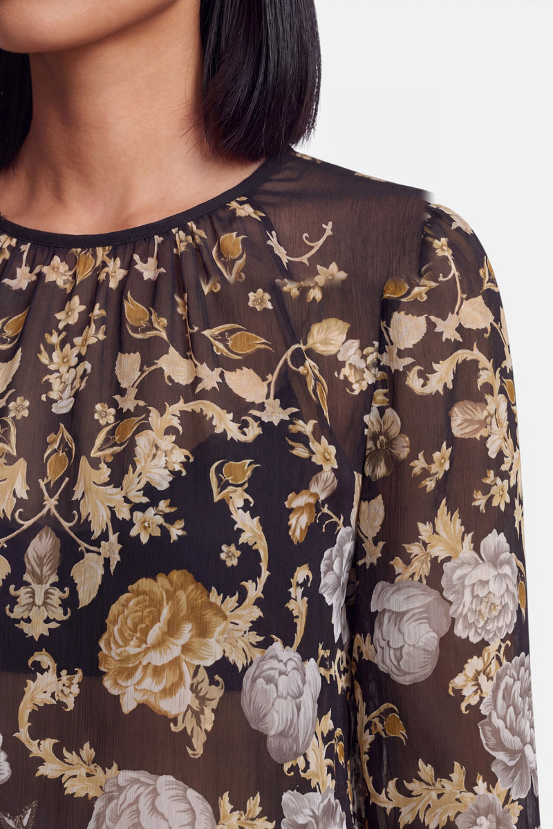 Eine Nahaufnahme des Oberkörpers einer Frau, die eine elegante Seidenbluse mit einem Blumenmuster in Beige-, Gold- und Weißtönen trägt. Die transparente Bluse ist am Ausschnitt gerafft und hat lange Ärmel. Ihr glattes, dunkles Haar ist zu sehen, und der Hintergrund ist schlicht weiß.