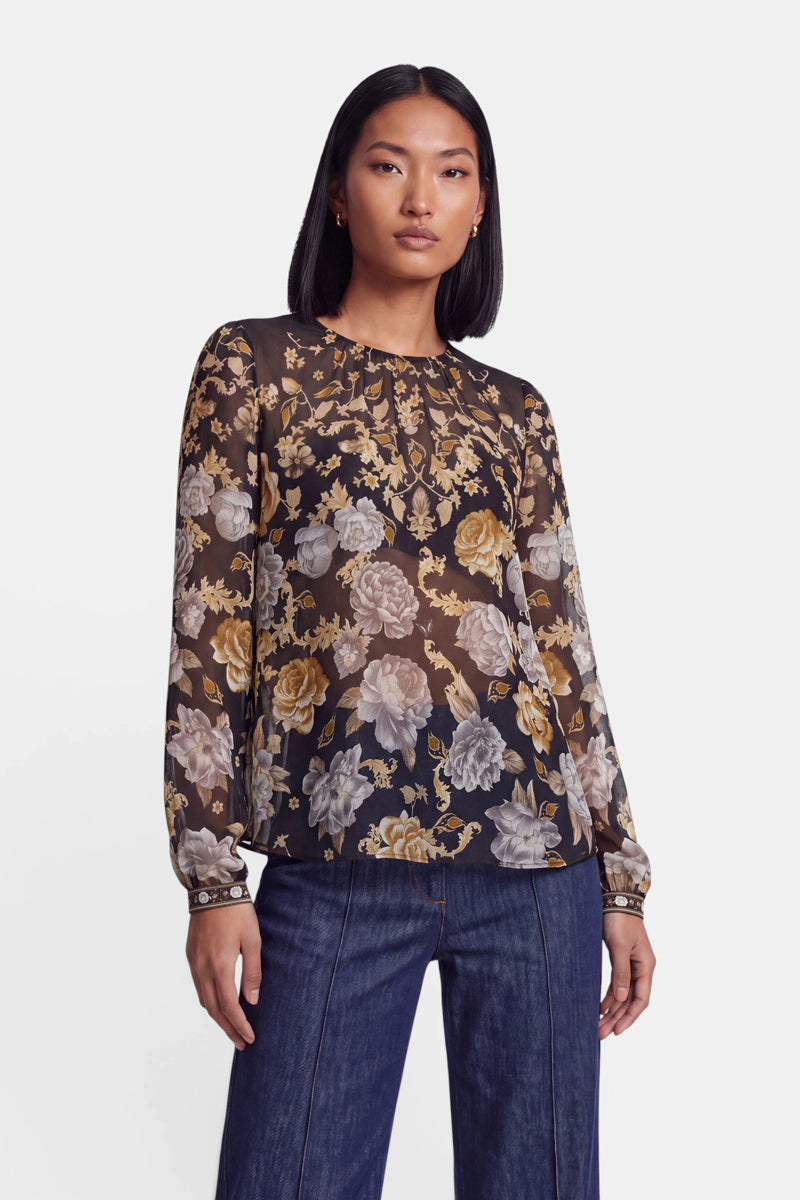 Eine Frau mit glattem, schulterlangem schwarzem Haar trägt eine transparente Bluse mit langen Ärmeln und einem gelb-cremefarbenen Blumenmuster, dazu eine blaue Jeans. Sie steht vor einem schlichten weißen Hintergrund und blickt mit ausdruckslosem Gesichtsausdruck nach vorne.