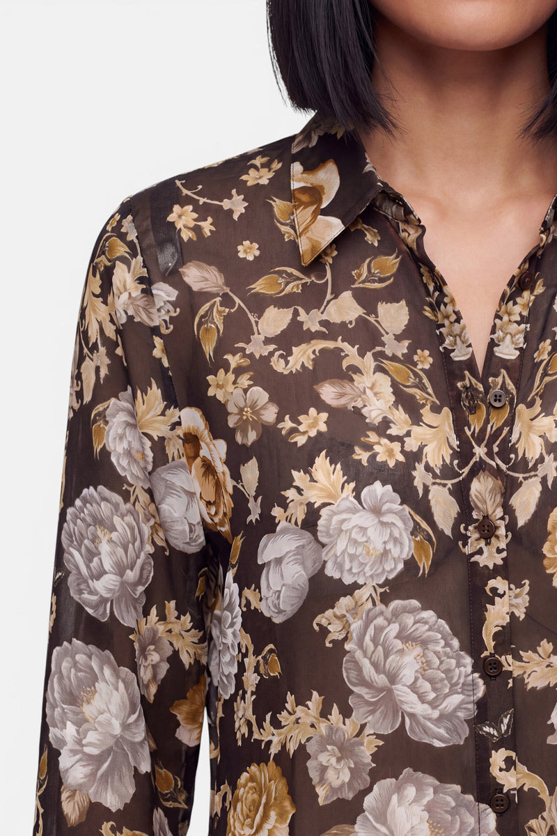 Eine Frau mit glattem, dunklem Haar trägt eine halbtransparente schwarze Seidenbluse mit einem Blumenmuster aus beigen, weißen und goldenen Blüten und Blättern. Die Hemdbluse hat einen Kragen und Knöpfe. Zu sehen sind nur ihr unterer Gesichtsbereich, ihr Hals, ihre Schultern und ihr Oberkörper.