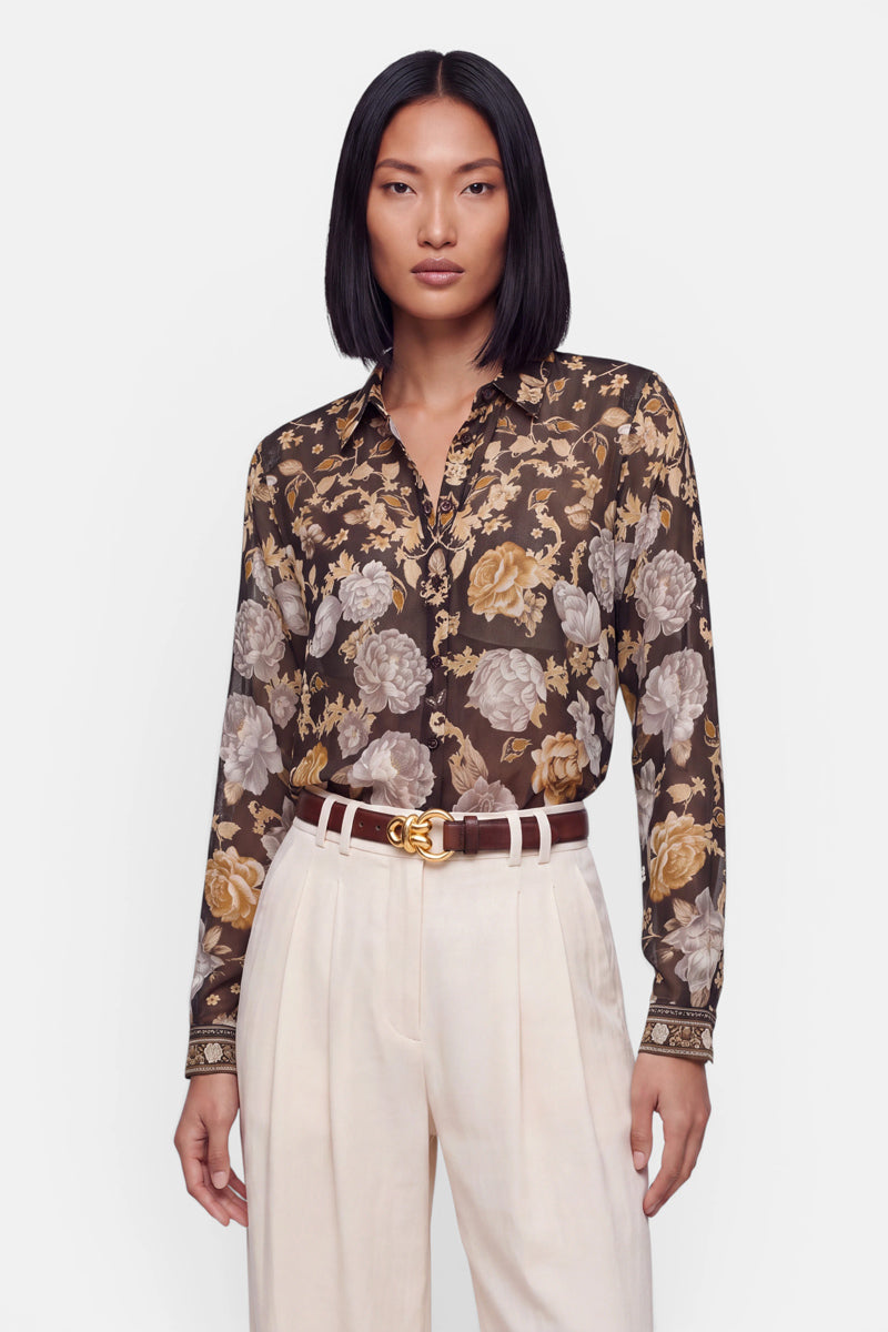 Eine Frau mit glattem schwarzem Haar trägt eine transparente Bluse mit einem Blumenmuster in Braun-, Gold- und Cremetönen, die sie in eine cremefarbene Hose mit hoher Taille, weitem Bein und Falten gesteckt hat. Außerdem trägt sie einen braunen Gürtel mit goldener Schnalle und hat einen neutralen Gesichtsausdruck.