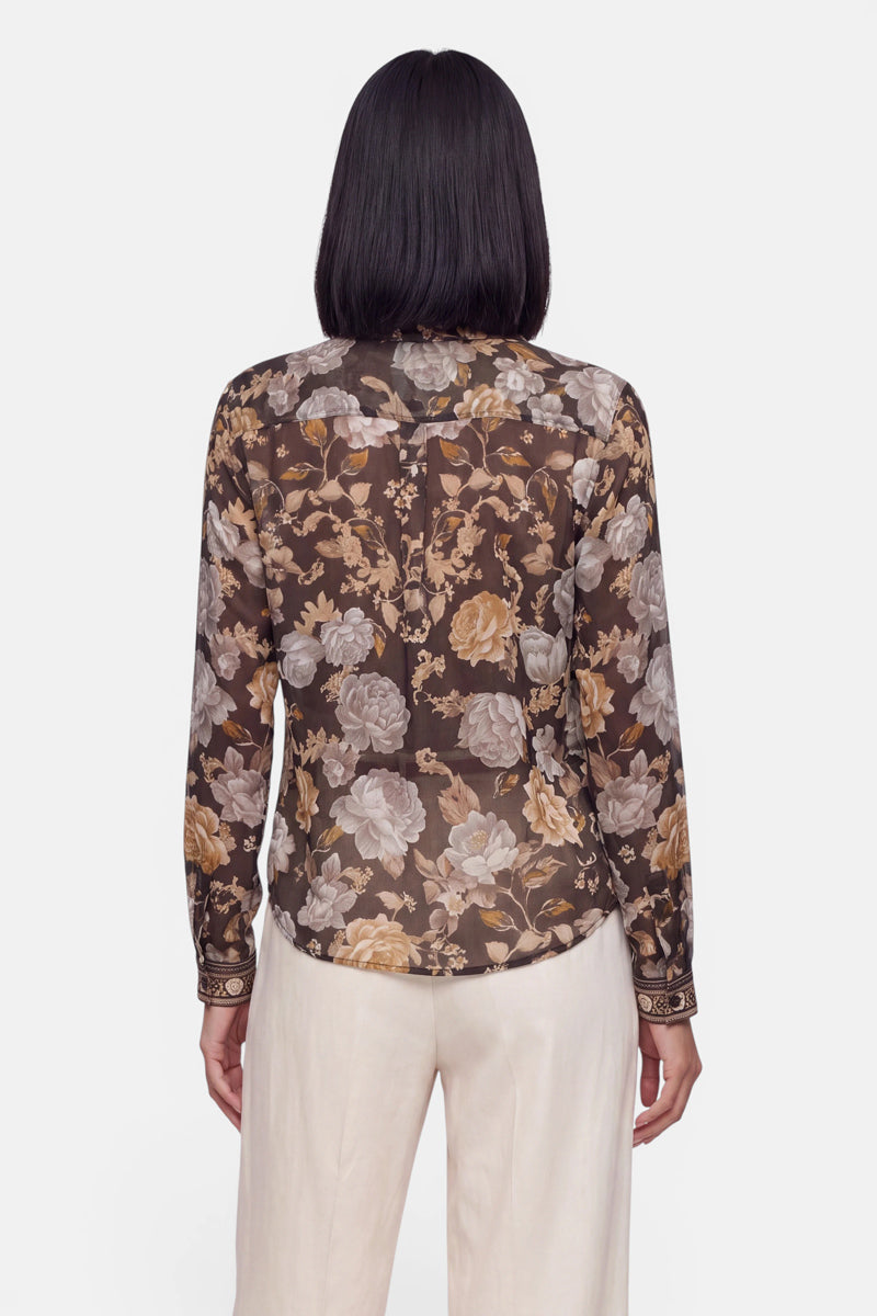 Eine Person mit glattem, dunklem Haar trägt eine transparente, langärmelige Seidenbluse mit einem Blumenmuster in Braun, Beige und Grau, dazu eine cremefarbene Hose. Die Aufnahme zeigt die Person von hinten vor einem schlichten weißen Hintergrund.