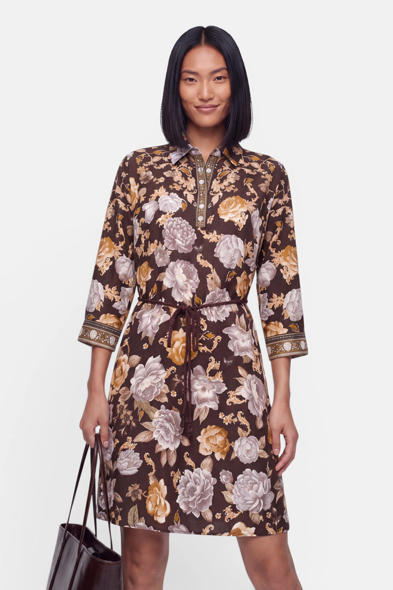 Eine Frau mit glattem schwarzem Haar trägt ein braunes Kleid mit Blumenmuster in Minilänge, mit Dreiviertelärmeln und aufwendigen Manschetten. Das ausgestellte Kleid hat eine Schleife an der Taille und ist mit beigen, goldenen und weißen Blumendetails verziert. Sie hält eine dunkle Tasche in einer Hand und lächelt sanft vor einem.