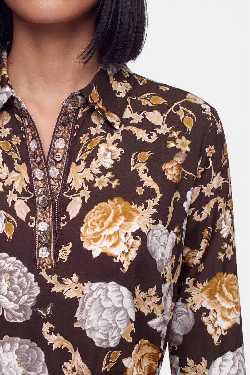 Eine Frau mit einem schwarzen Bob-Haarschnitt trägt ein dunkelbraunes Hemd mit Kragen, das mit aufwendigen floralen Mustern in Gold, Creme und Beige sowie dekorativen Knöpfen verziert ist und an ein opulentes Kleid mit Blumenprint erinnert. Vor einem schlichten weißen Hintergrund sind nur ihr Oberkörper und der untere Teil ihres Gesichts zu sehen.