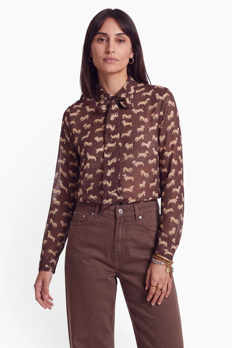 Vor einem weißen Hintergrund steht eine Frau, die eine braune Hemdbluse mit beigem Dackel-Print trägt, dazu eine braune Hose mit hoher Taille, goldene Creolen und Armbänder. Sie hat glattes, dunkles Haar und einen neutralen Gesichtsausdruck; eine Hand steckt in ihrer Hosentasche.