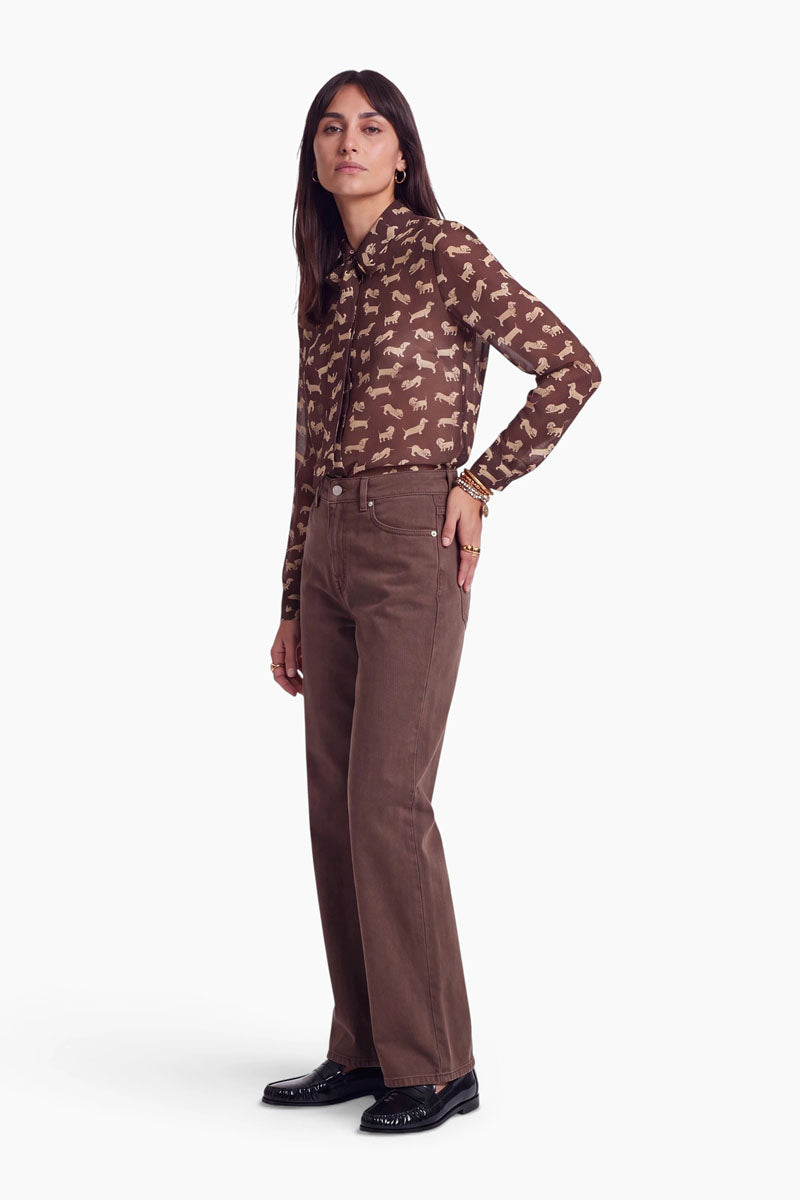 Vor einem weißen Hintergrund steht eine Frau, die eine braune, langärmelige Hemdbluse mit weißem Dackel-Aufdruck, eine braune Hose mit hoher Taille und weitem Bein, schwarze Schuhe sowie goldenen Schmuck trägt. Sie hat glattes, dunkles Haar und einen neutralen Gesichtsausdruck.
