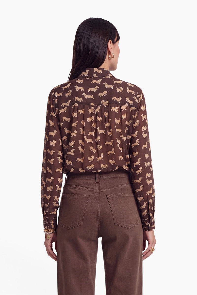 Eine Frau steht mit dem Rücken zur Kamera und trägt eine braune Hemdbluse mit beigem Tiermuster, die in eine braune Hose mit hoher Taille gesteckt ist. Sie hat langes dunkles Haar, goldene Creolen und goldene Ringe, während der Hintergrund schlicht weiß ist.