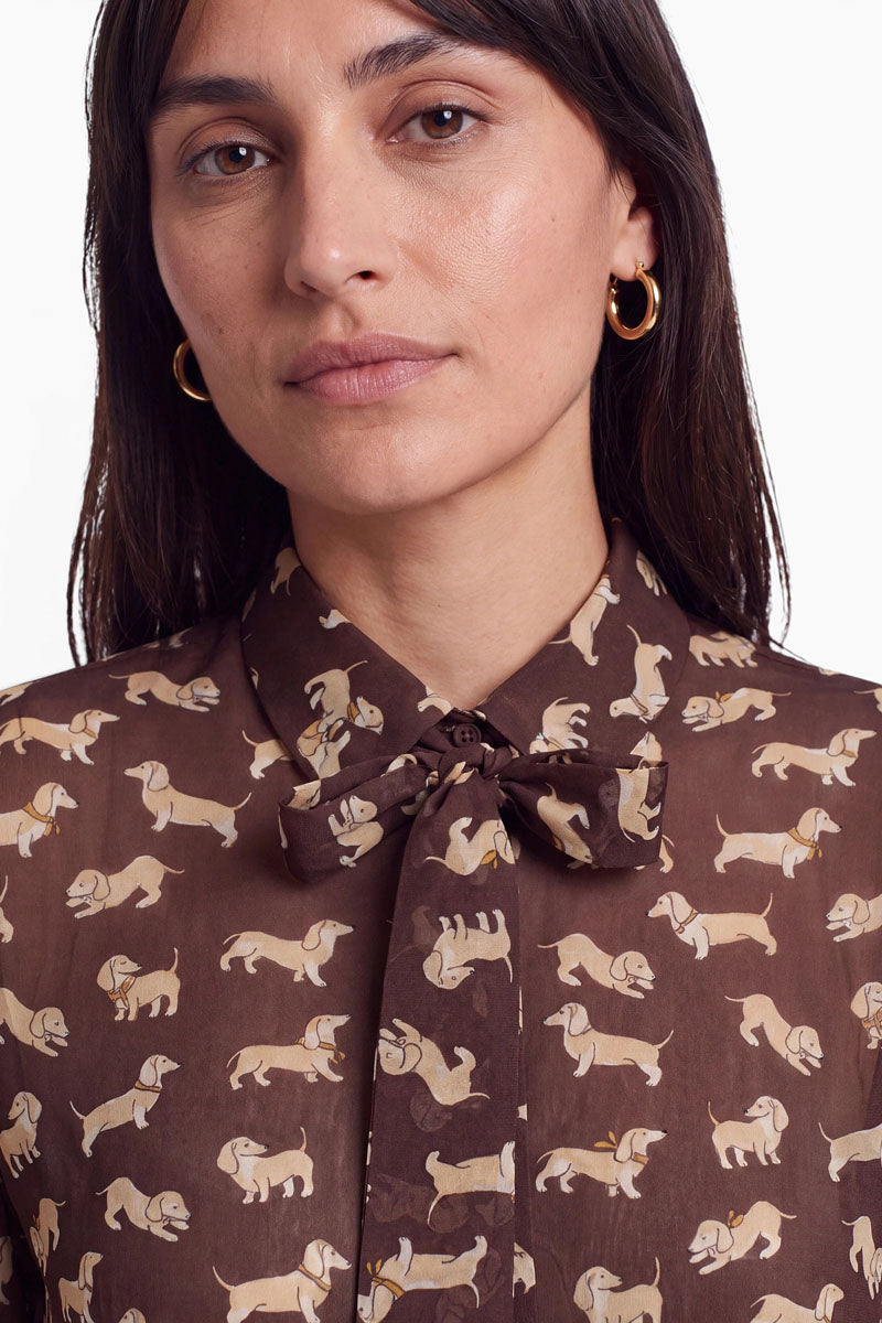 Eine Person mit glattem, dunklem Haar trägt goldene Creolen und eine braune Hemdbluse mit einem Muster aus beigen Dackeln. Die Hemdbluse ist am Kragen mit einer Schluppe verziert, auf der ein eleganter Dackel-Print zu sehen ist. Der Hintergrund ist schlicht weiß, und nur der Oberkörper sowie der untere Teil des Gesichts sind zu sehen.