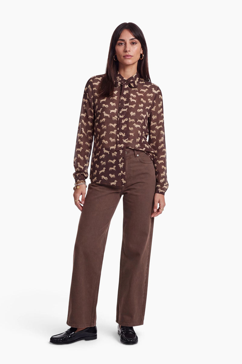 Die Frau steht mit dem Blick nach vorne und trägt eine braune Hemdbluse mit Pferdemotiven, eine braune Hose mit weitem Bein und schwarze Slipper. Sie hat glattes, dunkles Haar und trägt nur wenig Schmuck. Der Hintergrund ist schlicht weiß. Das Outfit wirkt lässig und zugleich stilvoll.