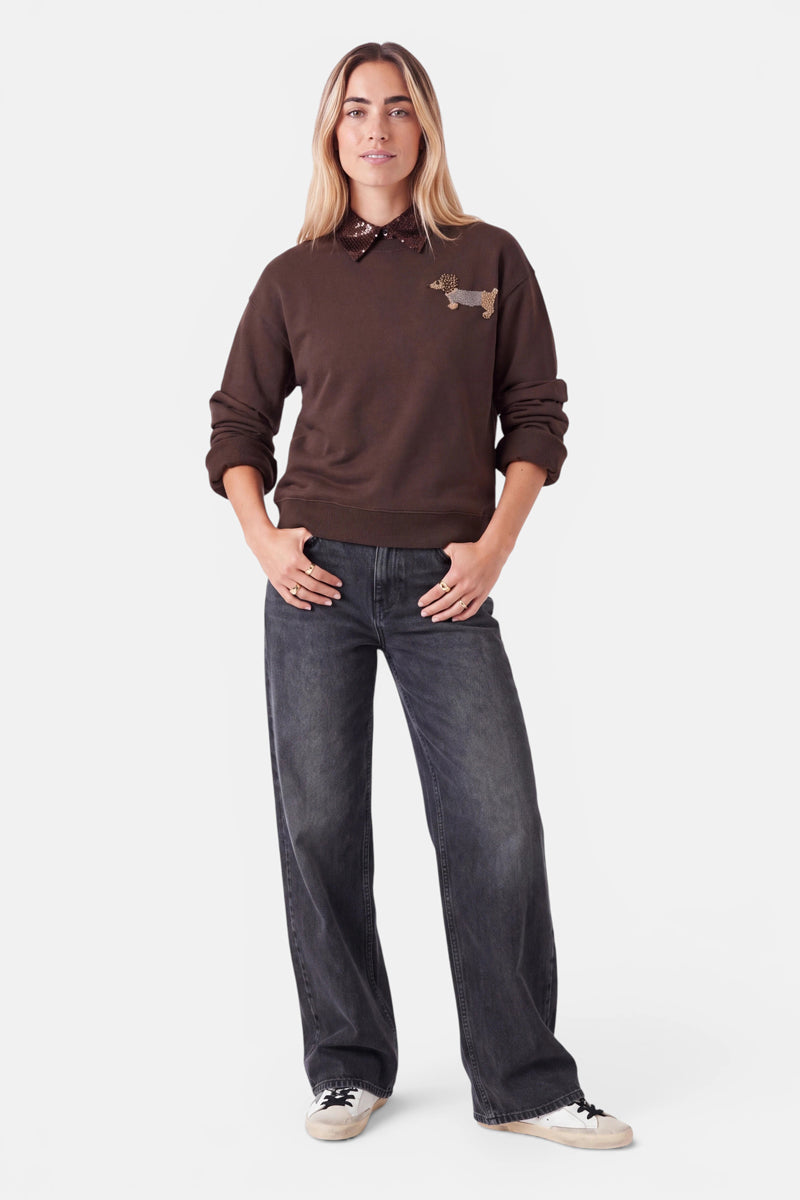 Eine Frau steht mit dem Blick nach vorne, die Hände in den Jeanstaschen. Sie setzt auf einen Blusen-Look mit lässigen Lagen und trägt einen braunen Pullover mit einem Dackel-Motiv auf der Brust, der über einem dunklen Hemd mit Kragen getragen wird, dessen Krageneinsatz am Ausschnitt hervorblitzt. Ihre dunkelblaue Jeans mit weitem Bein und weiße Sneaker runden das Outfit ab. Sie hat langes, glattes Haar und einen entspannten Gesichtsausdruck, während sie vor einem strahlend weißen Hintergrund steht.