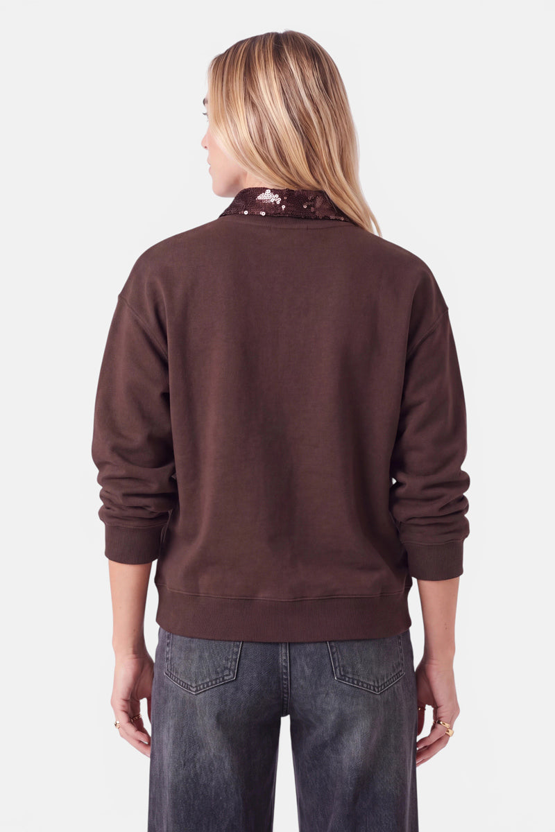 Eine Person mit langen blonden Haaren trägt ein braunes Sweatshirt mit einem glänzenden, paillettenartigen Krageneinsatz, der den Ausschnitt auflockert und einen dezenten Blusen-Look erzeugt. Die dunkelgraue Jeans rundet das Outfit ab. Die Person steht mit dem Rücken zur Kamera vor einem schlichten weißen Hintergrund, die Arme locker an den Seiten hängend.