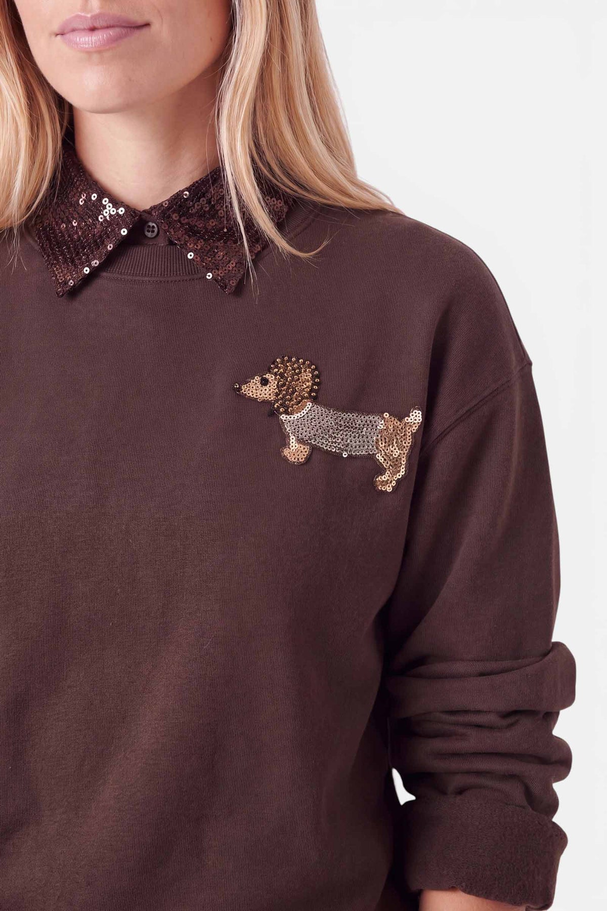 Eine Frau trägt einen braunen Pullover mit einem glänzenden Pailletten-Aufnäher in Form eines Dackels auf der Brust und einem dazu passenden braunen Paillettenkragen im charmanten Blusen-Look. Vor einem schlichten Hintergrund sind nur ihr unterer Gesichtsbereich und ihr Oberkörper zu sehen, wodurch der stilvolle Lagen-Effekt besonders gut zur Geltung kommt.